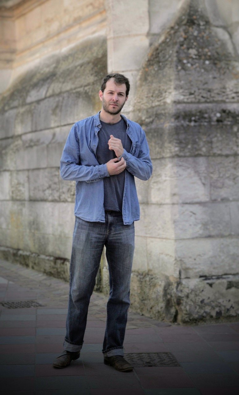 Portrait en pied, chemise bleue ouverte et jean, devant un mur de pierre