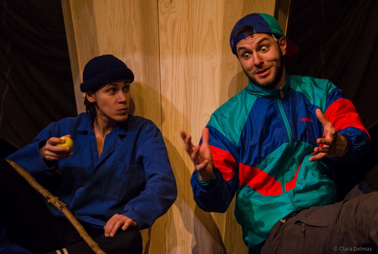 Deux comédiens devant un décor de bois, l’un en bleu de travail et bonnet, l’autre en casquette et veste de survêtement