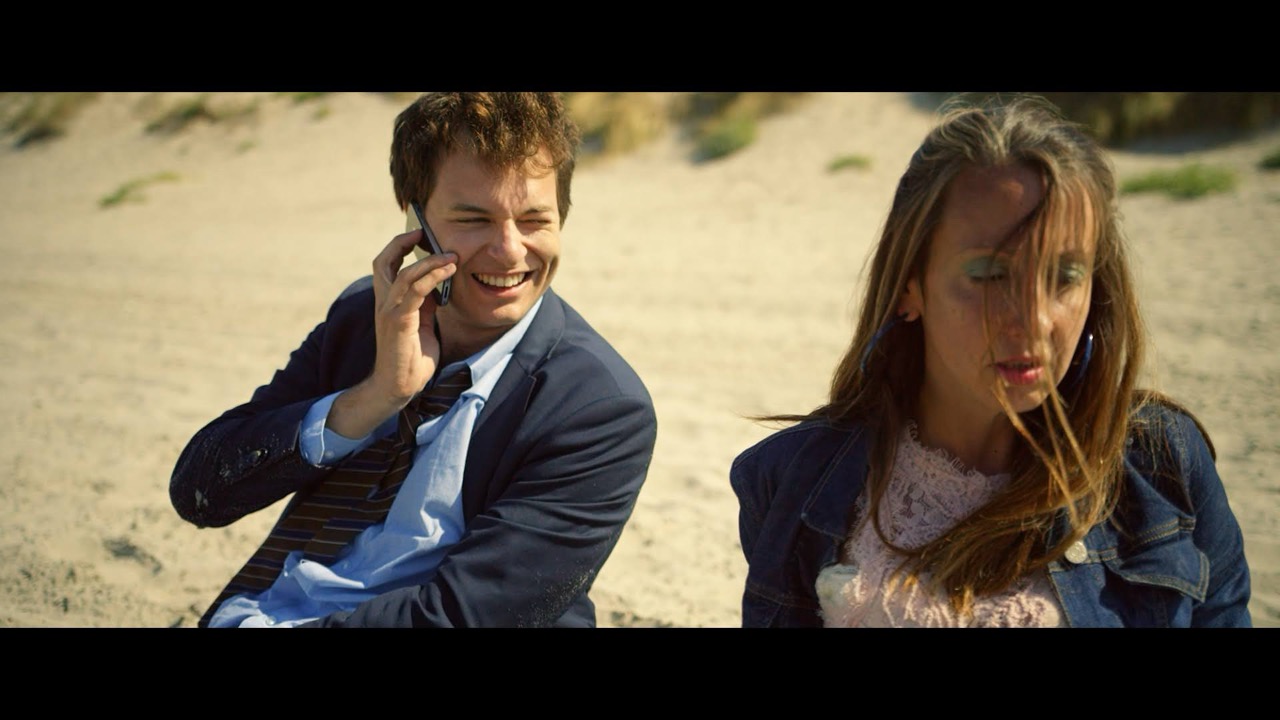 Image de film : sur une plage, un homme en costume rit au téléphone près d’une jeune femme aux cheveux au vent
