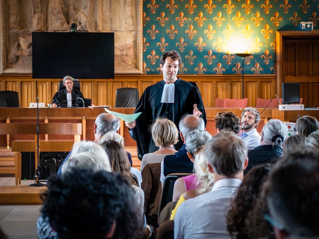 Dans une salle d’audience tapissée de fleurs de lys, un comédien en robe d’avocat plaide devant le public