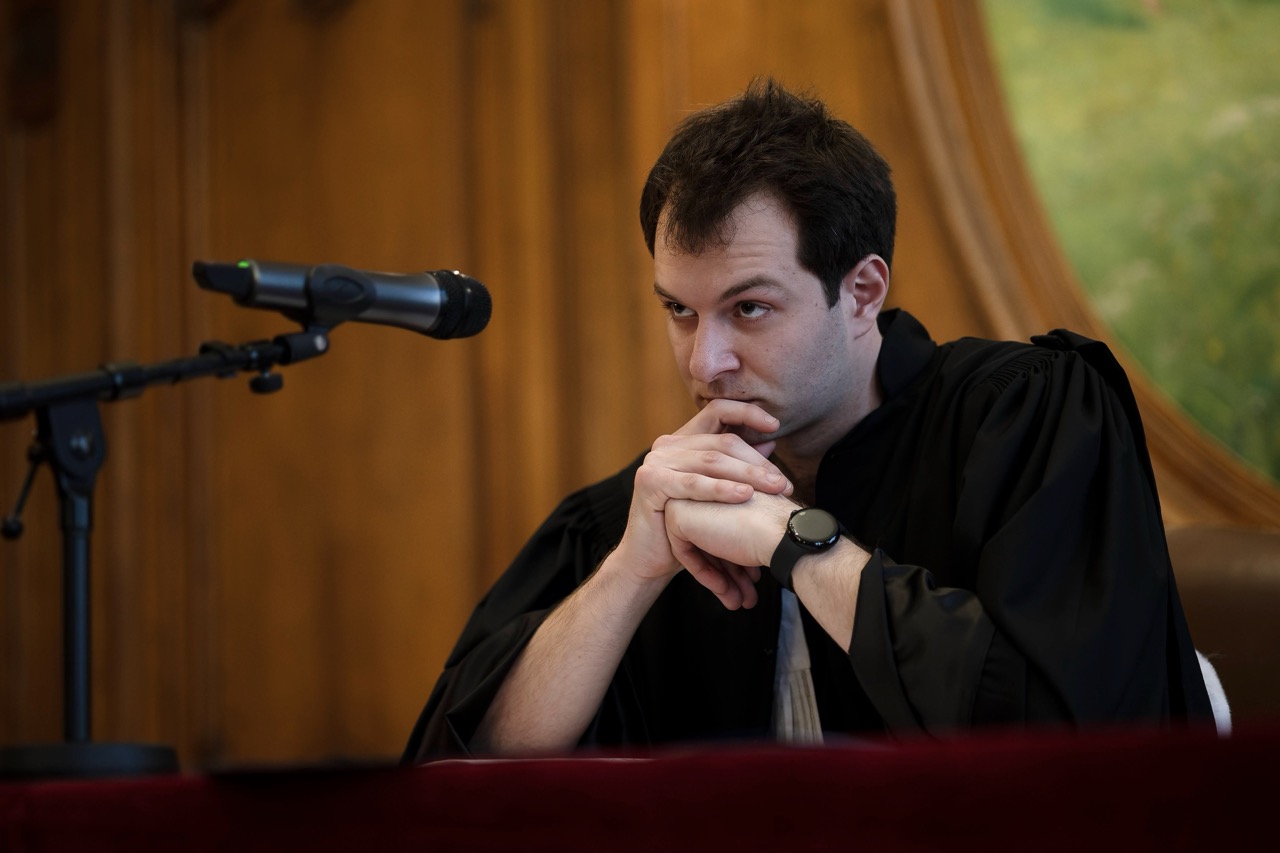 Salle d’audience lambrissée : en robe d’avocat, mains jointes devant un micro, l’air pensif