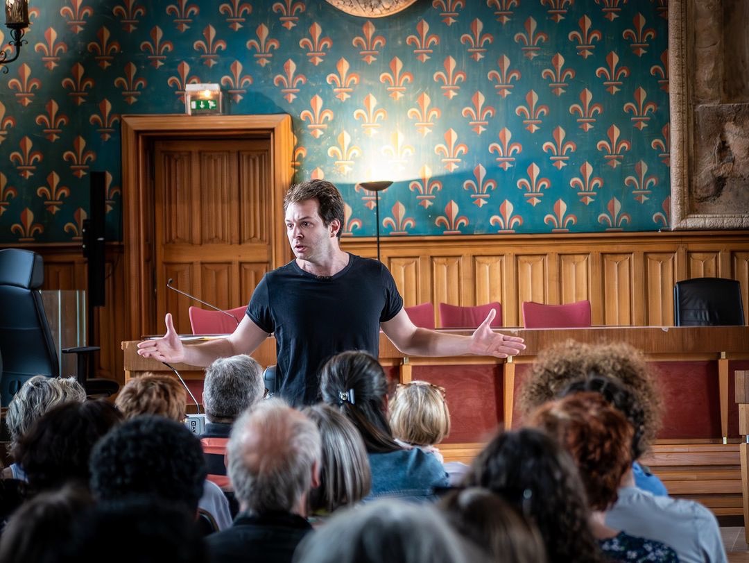 Même salle d’audience : un comédien en tee-shirt noir, bras ouverts, s’adresse au public
