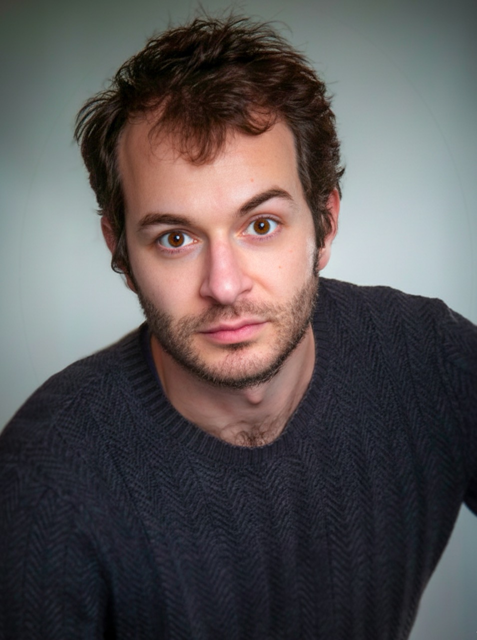 Portrait serré en pull gris, regard étonné vers l’objectif, sur fond gris clair